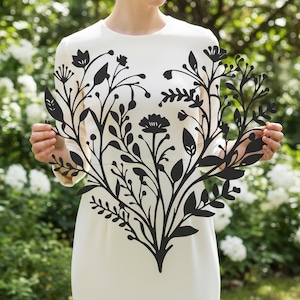 May include: A black metal floral wall art piece featuring a detailed design of leaves, flowers, and stems. The decorative piece is held in front of a person wearing a white dress, with a blurred green and white floral background.
