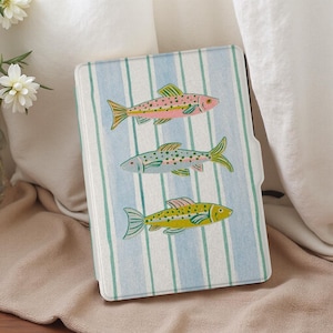 Puede incluir: Una funda rectangular con un diseño caprichoso de tres peces coloridos. Los peces son rosas, azules y amarillos, cada uno con patrones únicos, sobre un fondo de rayas verticales azules y blancas. La funda es probablemente para un dispositivo electrónico.
