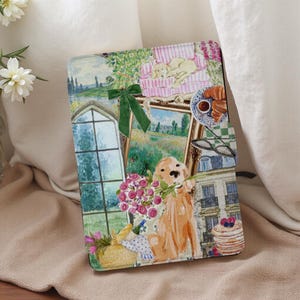 Peut inclure: Une housse de tablette décorative avec une illustration à l'aquarelle d'un golden retriever tenant des fleurs. Le design comprend une fenêtre, des œuvres d'art, des croissants et un chien endormi sur un canapé à rayures roses. Un nœud vert accentue le design.
