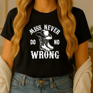 Op de afbeelding: Zwart T-shirt met de witte tekst "MISS NEVER DO NO WRONG" en een afbeelding van een cowboyhoed en -laarzen. De persoon draagt een blauwe spijkerbroek en heeft lang blond haar.
