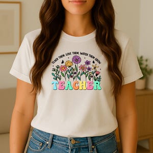 Op de afbeelding: Een crèmekleurig T-shirt met een kleurrijk bloemmotief en het woord "TEACHER" in een speels lettertype. De tekst "TEACH THEM. LOVE THEM. WATCH THEM GROW" staat in een boog boven de bloemen. Het ontwerp bevat vlinders en diverse bloemsoorten.