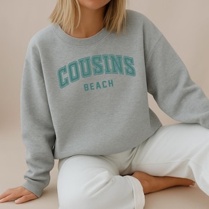 Peut inclure: Un sweat-shirt gris clair avec l'inscription "COUSINS BEACH" en lettres turquoise. Le sweat-shirt à col rond est porté avec un pantalon blanc. L'image évoque un style décontracté, idéal pour les loisirs.