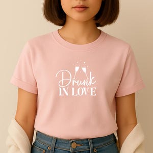 Op de afbeelding: Een lichtroze t-shirt met opgerolde mouwen. Het shirt toont de witte tekst "Drunk IN LOVE" met twee champagneglazen. Het model draagt een blauwe spijkerbroek.