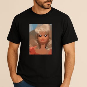Op de afbeelding: Zwart T-shirt met een pop-art afbeelding van een pop met wijd open ogen en een verbaasde uitdrukking. De pop heeft blond haar en staat tegen een lichtblauwe achtergrond. Het shirt heeft een klassieke ronde hals.