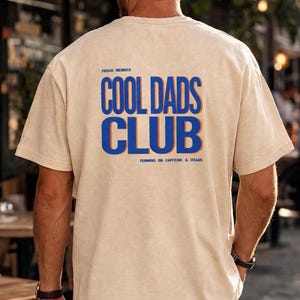 Può includere: T-shirt beige con la scritta "COOL DADS CLUB" in blu e arancione, con "PROUD MEMBER" sopra e "RUNNING ON CAFFEINE & CHAOS" sotto. La maglietta è indossata da una persona vista da dietro.