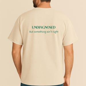 Op de afbeelding: Beige t-shirt met de groene tekst "UNDIAGNOSED" en de zin "But something ain't right". Het shirt heeft korte mouwen en is gemaakt van een zacht materiaal.
