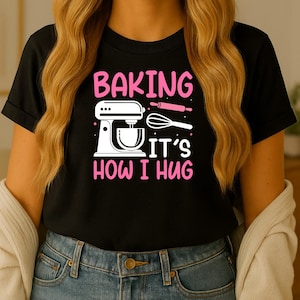 Op de afbeelding: Zwart T-shirt met een roze en witte opdruk met de tekst "BAKING IT'S HOW I HUG". De afbeelding bevat een keukenmixer, deegroller en garde. Het T-shirt wordt gedragen door een persoon met lang blond haar.