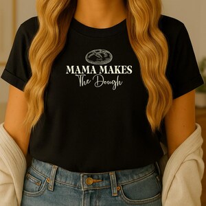 Op de afbeelding: Zwart T-shirt met de tekst "MAMA MAKES The Dough" in witte letters en een broodafbeelding. Het shirt heeft een ronde hals en wordt gedragen door een persoon met lang blond haar.