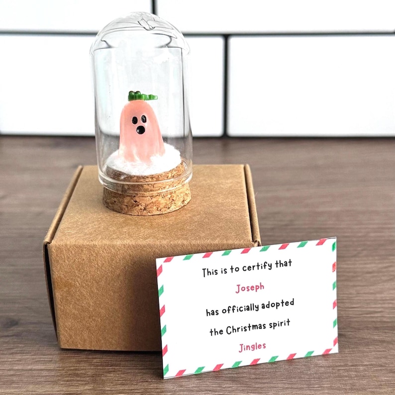 Mini pink Christmas ghost figurine named Jingles in a glass dome with snow and gold stars, displayed on a black gift box next to a certificate card that says “Sophie has adopted the Christmas spirit Jingles.” Cute and unique holiday gift, Christmas decor, or stocking stuffer with festive background and pine branch.