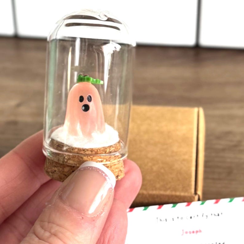 Mini pink Christmas ghost figurine named Jingles in a glass dome with snow and gold stars, displayed on a black gift box next to a certificate card that says “Sophie has adopted the Christmas spirit Jingles.” Cute and unique holiday gift, Christmas decor, or stocking stuffer with festive background and pine branch.
