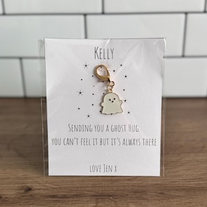 Könnte beinhalten: Ein weißer Geisteranhänger mit einem goldenen Verschluss, der an einer Karte befestigt ist. Auf der Karte steht oben "KELLY" und darunter "SENDING YOU A GHOST HUG. YOU CAN'T FEEL IT BUT IT'S ALWAYS THERE LOVE JEN X."