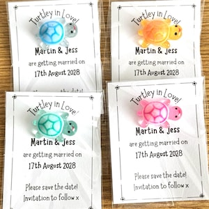 May include: Four wedding favors featuring turtle designs in blue, orange, green, and pink. Each favor includes a card with the text "Turtley in Love," the names "Martin & Jess," and the wedding date, August 17th, 2028. The cards also say "Please save the date!"