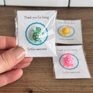 Puede incluir: Pequeños pines con forma de tortuga en diferentes colores, empaquetados individualmente. Cada tarjeta blanca lleva el texto "Thank you for being" y "Turtley awesome!" en negro, con un círculo azul alrededor de la tortuga.