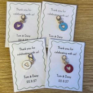 May include: Four cards with charms, each featuring a heart in a different colour: purple, blue, white, and red. The cards read "Thank you for celebrating with us!" and "Tom & Daisy 22.8.27". The charms have a gold clasp.