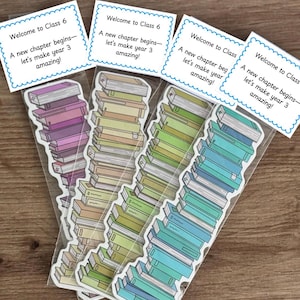 Puede incluir: Cuatro paquetes transparentes con pegatinas con forma de libro en varios colores: morado, beige, verde y azul. Cada paquete tiene una tarjeta blanca con el texto "Welcome to Class 6" y el mensaje "A new chapter begins - let's make year 3 amazing!"