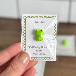Puede incluir: Un pequeño pin en forma de rana verde brillante sobre una tarjeta blanca con el texto "You are TOADally Brilliant! From Miss Jones". La tarjeta tiene un borde festoneado verde y está en un envoltorio de plástico transparente.