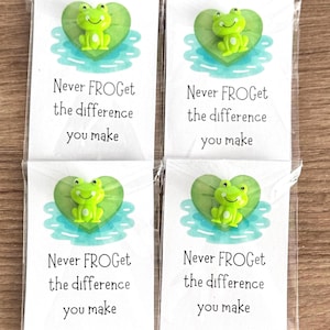 May include: Four identical white rectangular cards in clear plastic sleeves, each featuring a small green frog figurine on a green heart-shaped lily pad illustration. The text on each card reads: Never FROGet the difference you make.