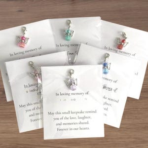 Könnte beinhalten: Eine Sammlung von Engelscharms auf weißen Karten. Jeder Engelscharm hat eine unterschiedlich farbige Perle, darunter rosa, grün, rot, blau und lila. Die Karten tragen den Text "In loving memory of" und eine Botschaft zum Gedenken an geliebte Menschen.
