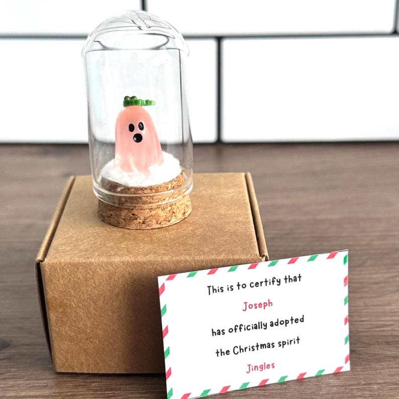 Mini pink Christmas ghost figurine named Jingles in a glass dome with snow and gold stars, displayed on a black gift box next to a certificate card that says “Sophie has adopted the Christmas spirit Jingles.” Cute and unique holiday gift, Christmas decor, or stocking stuffer with festive background and pine branch.