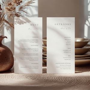 Pode incluir: Dois cartões de menu de casamento bege com os nomes "Anne & Tim" em uma escrita delicada. Os cartões listam itens do menu e bebidas. O fundo inclui um vaso, plantas secas e pratos empilhados, criando uma atmosfera acolhedora.