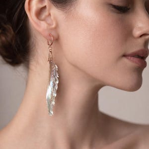 Pode incluir: Brincos de argola em ouro rosa com um pendente pendurado em forma de pena iridescente. O pendente tem um design texturizado e em camadas, que capta a luz. Os brincos são um acessório delicado.
