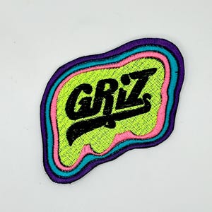May include: Embroidered patch with the word "GRIZ" in black, set against a neon green background. The patch has a unique, irregular shape with a pink, teal, and purple border. The patch is likely for clothing or accessories.