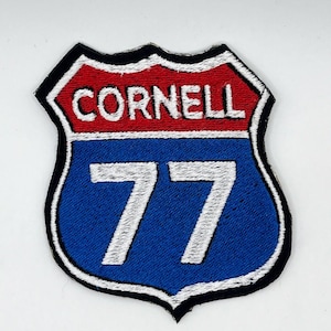 May include: Embroidered patch in the shape of a road sign. The top section is red with the word "CORNELL" in white. The bottom section is blue with the number "77" in white. The patch has a black border.