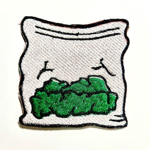 May include: Embroidered patch featuring a white bag with a black outline, filled with green, leafy material. The bag has a textured appearance and is sealed with black lines. The patch is designed to be ironed on or sewn onto fabric.