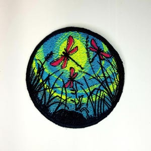 May include: A round embroidered patch with a blue background and a yellow moon. Three red dragonflies are flying in front of the moon. The patch is surrounded by black grass.