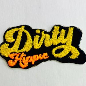 May include: Embroidered patch with the words "Dirty Hippie" in yellow and orange thread on a black background. The patch has a unique, irregular shape and is likely designed to be sewn onto clothing or accessories.
