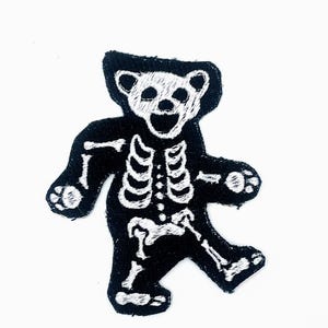 May include: Black and white embroidered patch in the shape of a dancing bear skeleton. The bear has a black outline with white embroidered details depicting the skeleton. The bear is in a dancing pose with its arms and legs spread out.