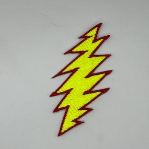 May include: Embroidered patch in the shape of a yellow lightning bolt with a red border. The patch is on a white background. The lightning bolt design is a zig-zag shape.