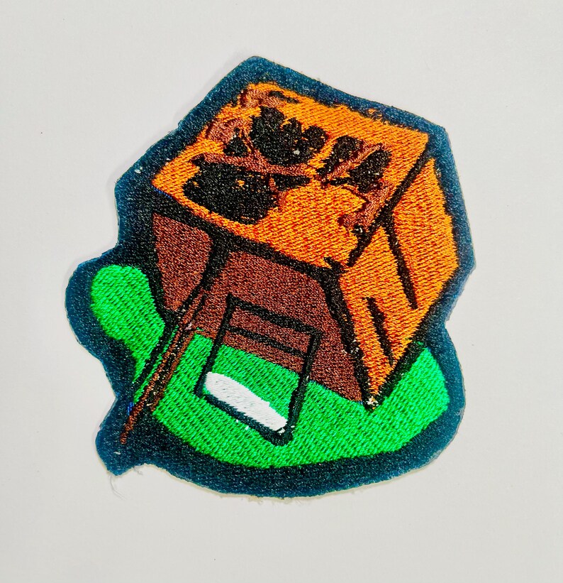 May include: Embroidered patch featuring a stylized brown and orange cube with a green base. The cube has a dark brown roof and a white and green window. The patch is outlined in dark blue.