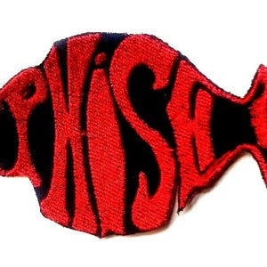 May include: A red and black embroidered patch in the shape of a fish. The word "PHISH" is spelled out in a stylized font, with the letters forming the body of the fish. The patch has a white background.