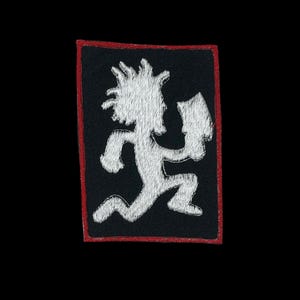 May include: Embroidered patch featuring a white figure with wild hair and a hatchet, running to the right. The design is on a black rectangular background with a red border. The patch is likely for clothing or accessories.