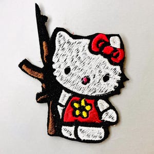 K&ouml;nnte beinhalten: Wei&szlig;er gestickter B&uuml;geleisen-Aufn&auml;her von Hello Kitty, die ein braunes Gewehr mit einer roten Schleife und einem roten Shirt mit einer gelben Blume h&auml;lt.