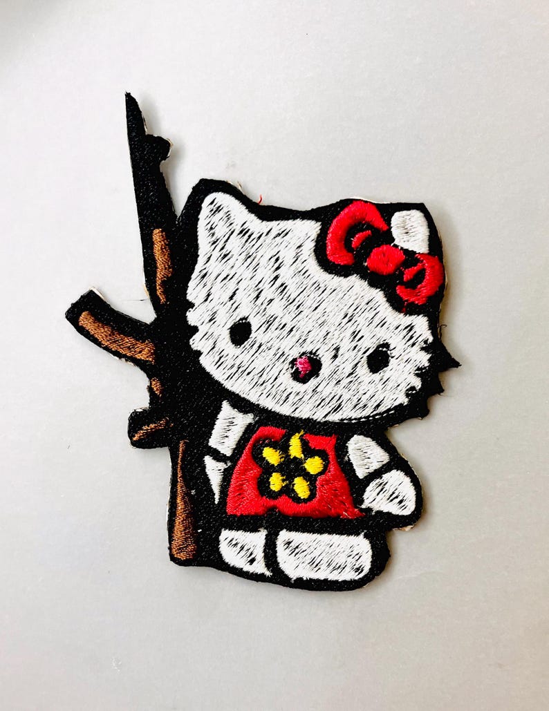 K&ouml;nnte beinhalten: Wei&szlig;er gestickter B&uuml;geleisen-Aufn&auml;her von Hello Kitty, die ein braunes Gewehr mit einer roten Schleife und einem roten Shirt mit einer gelben Blume h&auml;lt.