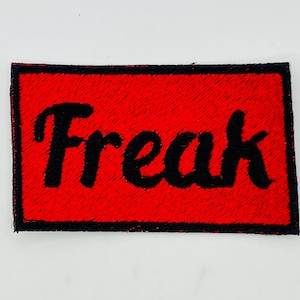 May include: A rectangular patch with a red background and the word "Freak" in black, cursive lettering. The patch has a black border and is likely an iron-on or sew-on patch for clothing or accessories.