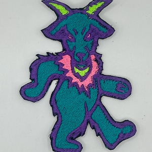 May include: Embroidered patch featuring a stylized bear-like creature. The patch has a teal body, purple outline, and accents of pink and neon green. The bear is depicted with a stern expression and a jagged collar.