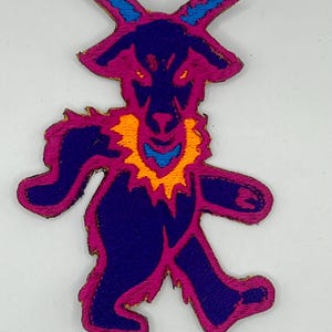 May include: Embroidered patch featuring a stylized goat-like creature in shades of purple, blue, and orange. The patch has a textured appearance and a gold border. The design is a whimsical representation of a bear.