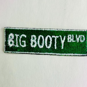 May include: Embroidered patch in the shape of a street sign. The patch is green with white embroidered text that reads "BIG BOOTY BLVD". The patch has a white border and is likely designed to be sewn onto clothing or accessories.