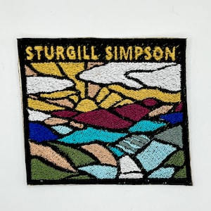 May include: Embroidered patch featuring the text "STURGILL SIMPSON" in yellow on a black background. The patch depicts a colorful landscape with a sun, clouds, and various geometric shapes in shades of yellow, red, blue, and green.