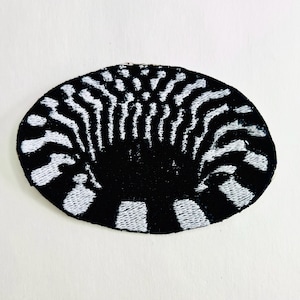 May include: Black and white embroidered patch with a unique design. The patch features a black base with white, curved lines radiating from the center, creating a textured, eye-catching pattern. The patch is oval-shaped.