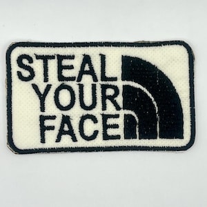 May include: A rectangular patch with the words "STEAL YOUR FACE" in black block letters on a cream-colored background. The right side features a black, stylized arch design, all framed by a black border.