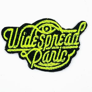 Op de afbeelding: Een zwart embleem met de tekst "Widespread Panic" in neon groen, met een gestileerd oogsymbool. Het lettertype is opvallend. Het embleem is waarschijnlijk voor kleding of accessoires en heeft een getextureerde afwerking.