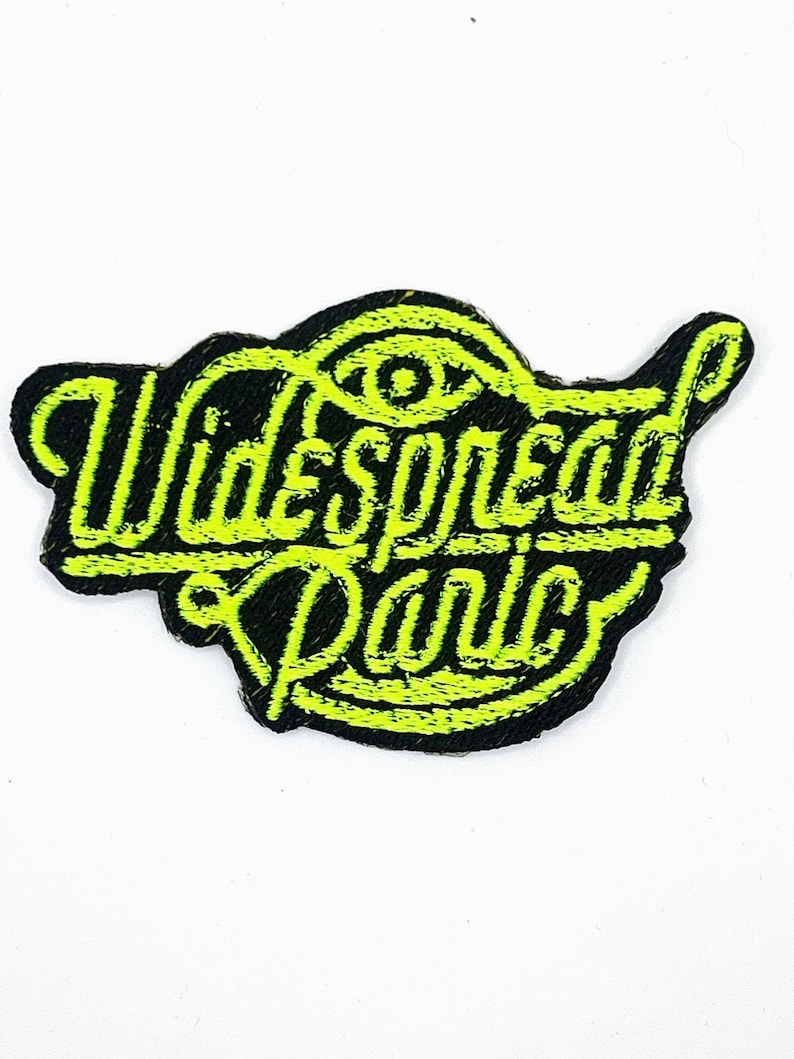 Op de afbeelding: Een zwart embleem met de tekst "Widespread Panic" in neon groen, met een gestileerd oogsymbool. Het lettertype is opvallend. Het embleem is waarschijnlijk voor kleding of accessoires en heeft een getextureerde afwerking.
