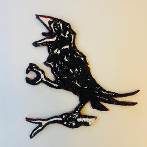 May include: Black embroidered iron-on patch of a crow with its wings spread out. The crow is perched on a branch with its beak open.