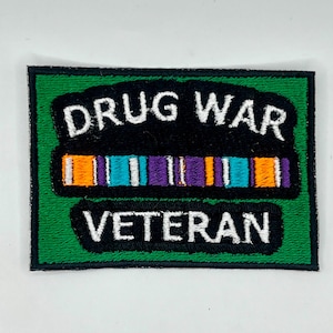 May include: Embroidered patch with the words "DRUG WAR VETERAN" in white on a black background. The patch features a colorful ribbon design with orange, teal, and purple stripes, set against a green backdrop.