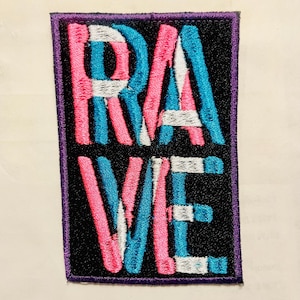 May include: Embroidered patch with the word "RAVE" in pink, blue, and white letters on a black background. The patch has a purple border. The letters are stylized and bold.