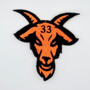 May include: Embroidered patch featuring an orange goat head with black accents and the number 33. The patch has a detailed design with a textured appearance, suitable for application on clothing or accessories.
